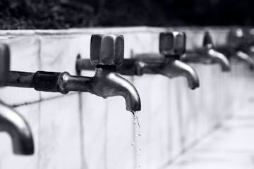 Black and white image of a row of faucets with water dripping, highlighting plumbing details.